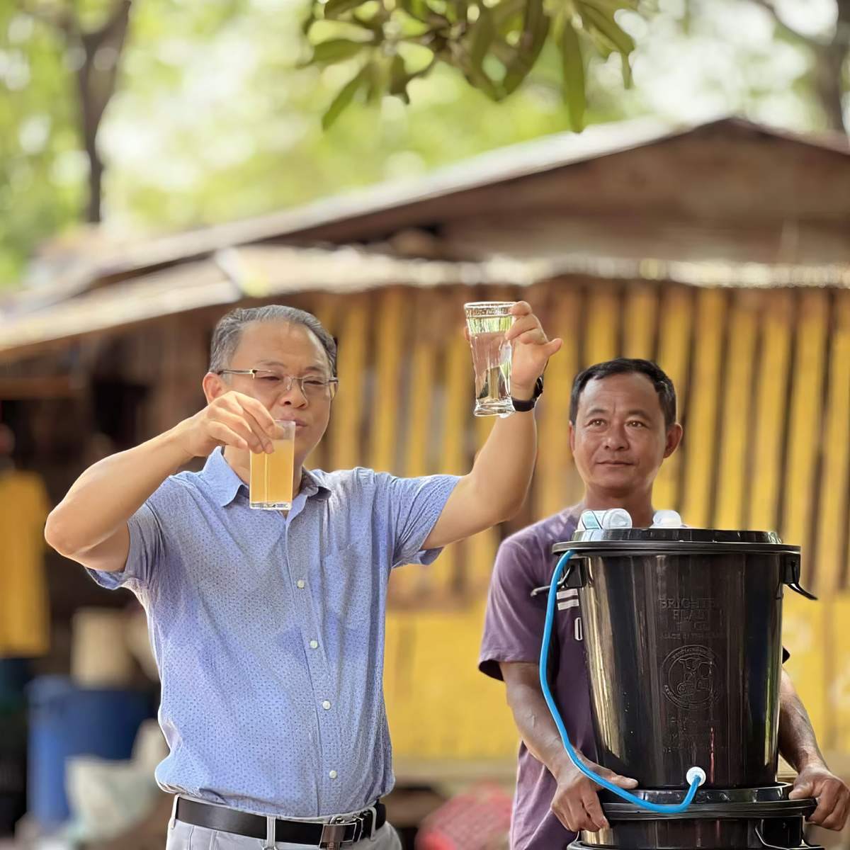 A man raises a cup of filtered water beside a filter bucket