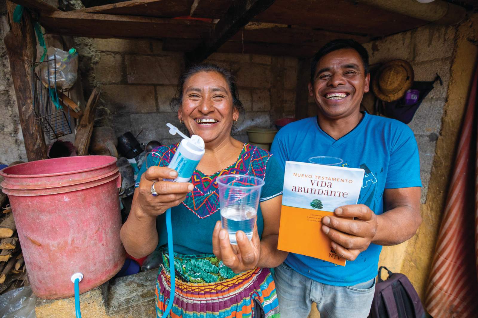 A smiling couple holds up their filter, a cup of clean water, and a Gospel booklet